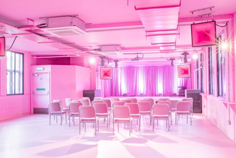 A brightly lit event space in Manchester, awash with pink lighting, featuring rows of empty chairs facing a stage with purple curtains, ready for an audience to arrive.