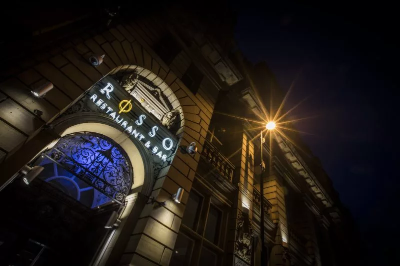 An illuminated sign for "Rosso Restaurant & Bar" mounted on the facade of an elegant building under a night sky, with a radiant street light enhancing the festival atmosphere by creating a starburst effect above
