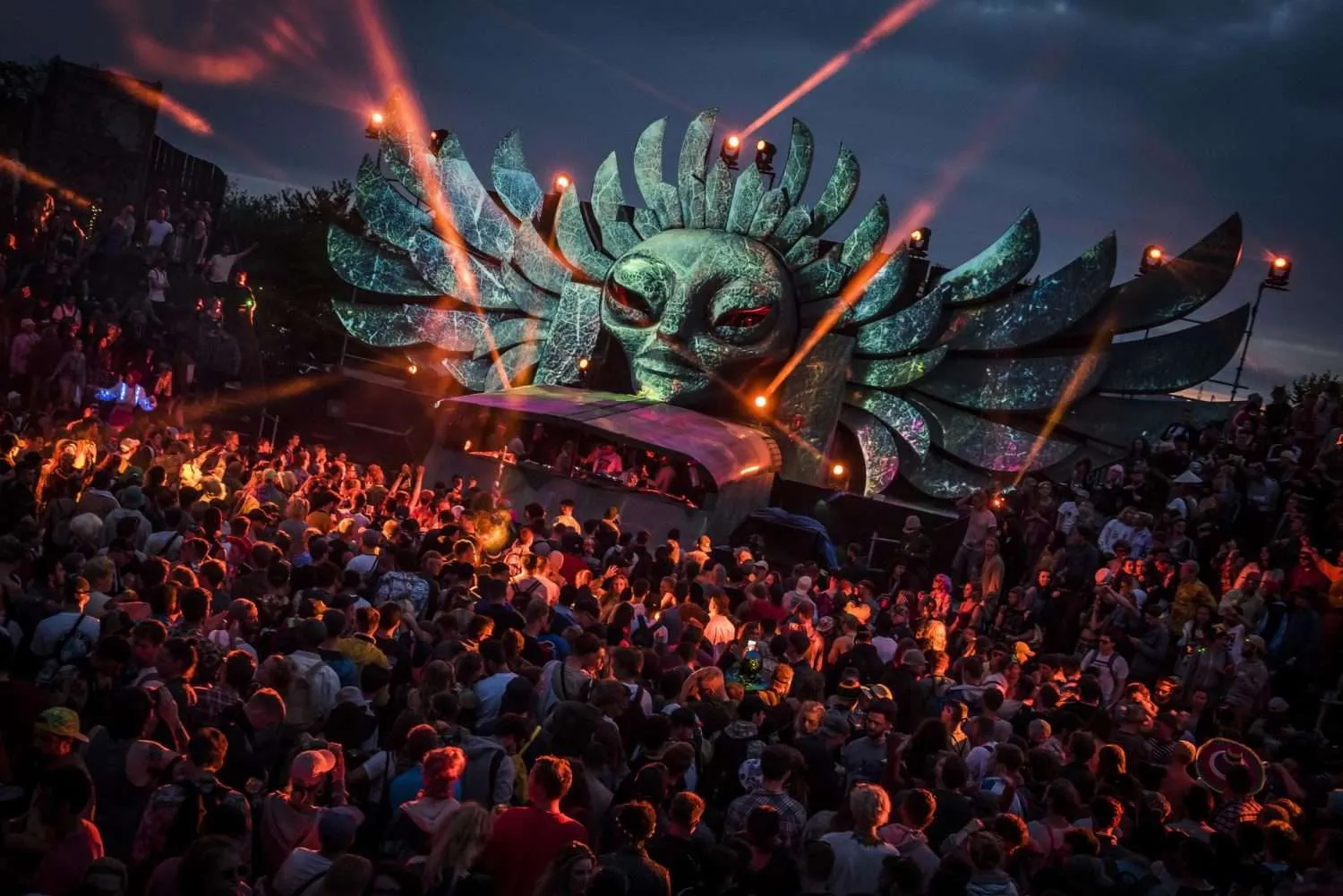 A vibrant outdoor music festival scene at dusk, featuring a large, illuminated stage with an extravagant alien-themed design and state-of-the-art festival audio production, surrounded by an energetic crowd of festival-goers immersed