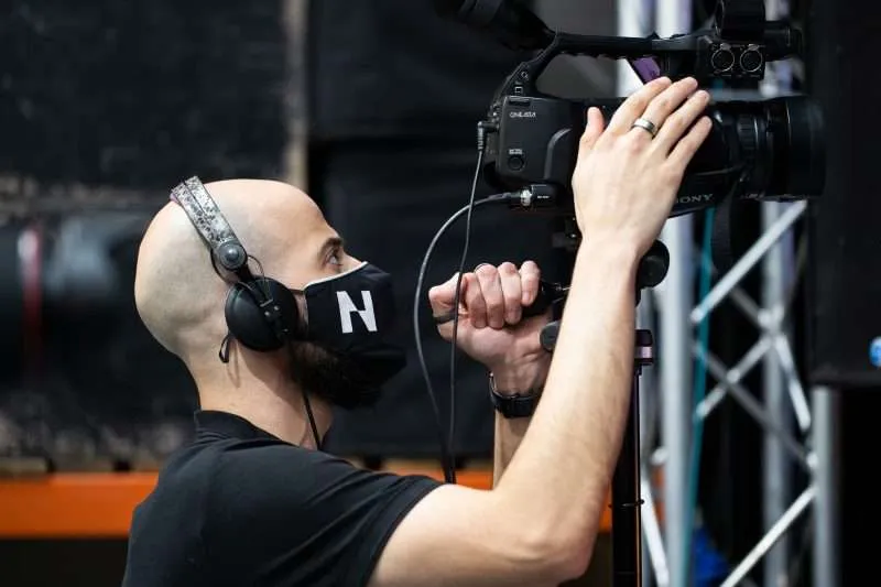 A cameraman wearing a mask and headphones operates a professional video camera in a studio, focusing intently on capturing the scene for a virtual event.
