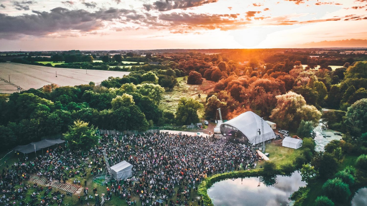 An aerial view of an outdoor concert, featuring advanced audio production, nestled in lush greenery, with a large crowd gathered by the stage as the sun sets on the horizon.