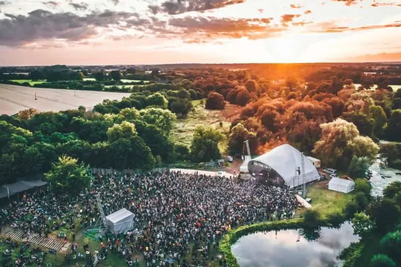 A vibrant outdoor concert at sunset, with lively audio production filling the air as a crowd gathers around the stage amidst a picturesque landscape of trees and a tranquil pond.