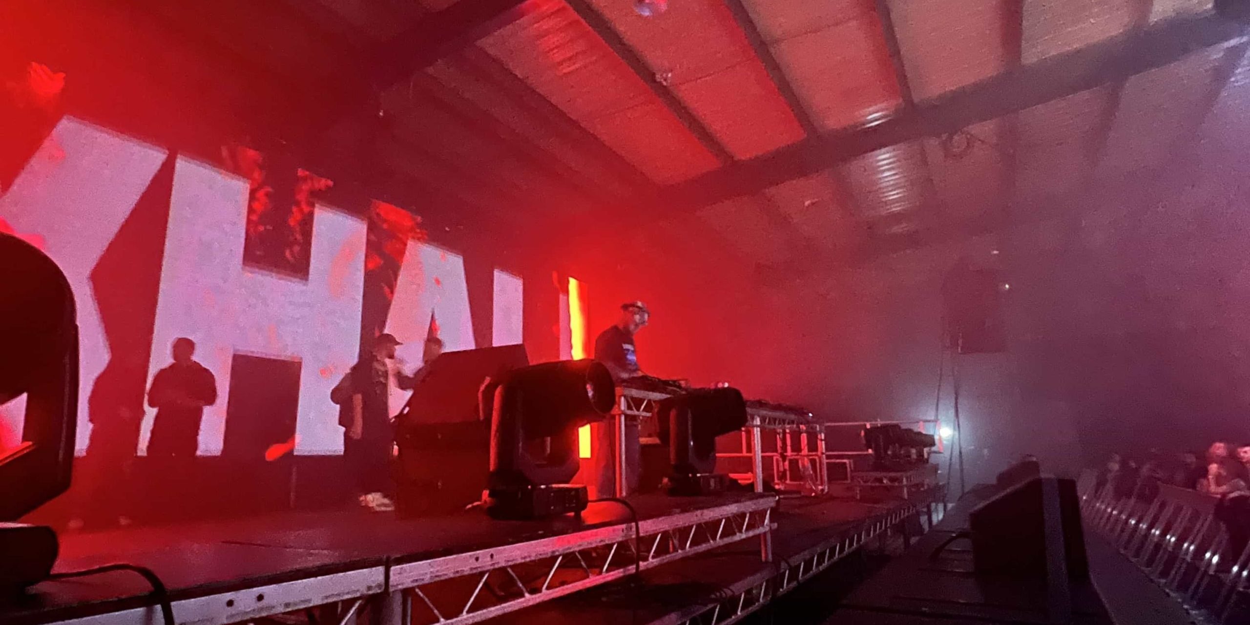 A moody concert setting with vivid red lighting hire and silhouettes of people, with the word 