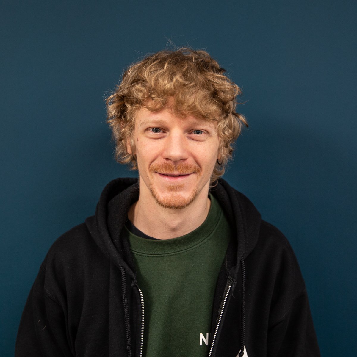 A smiling man with curly blond hair and a relaxed demeanor, wearing a green AV company logo shirt under a black zip-up hoodie, set against a blue background.