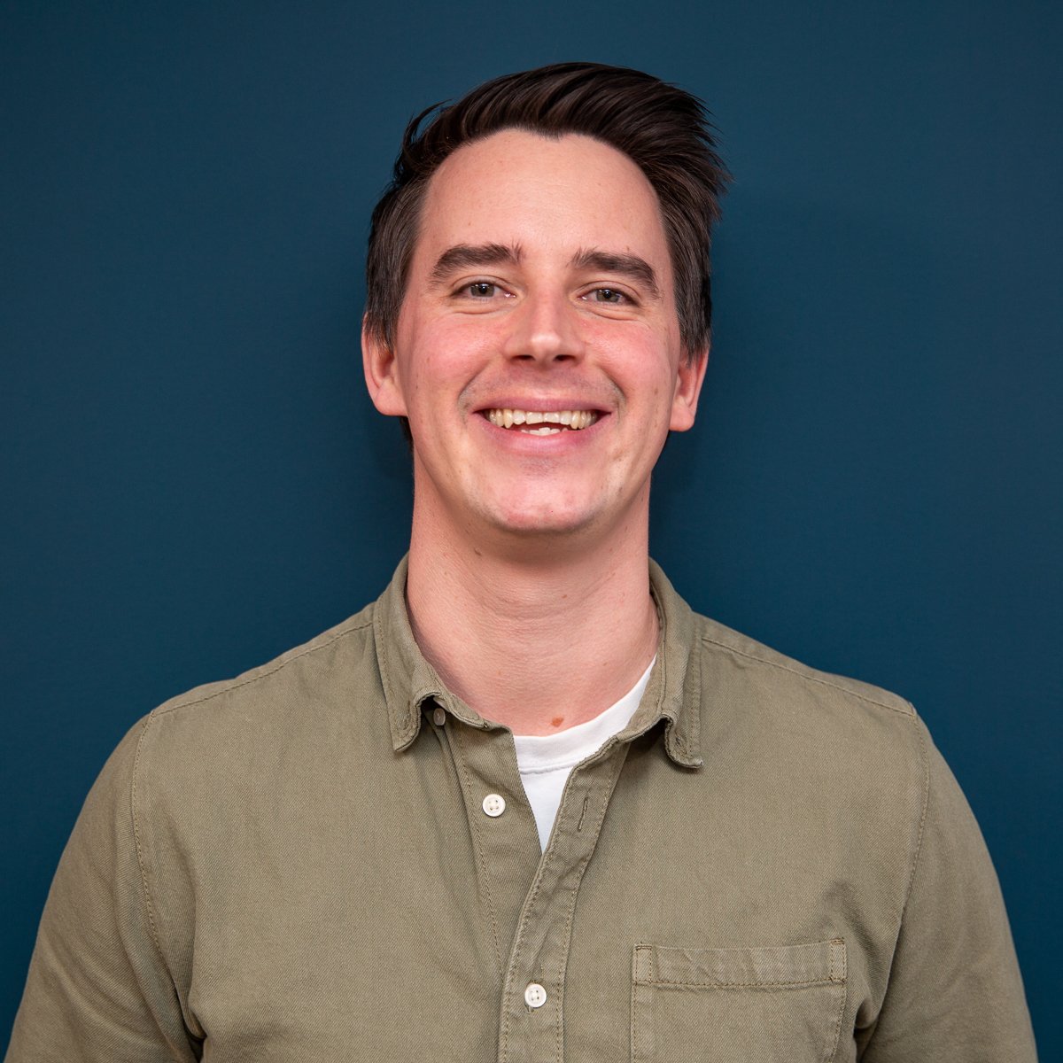 A cheerful man with a friendly smile wearing a casual olive green shirt against a deep blue background, representing an AV company.