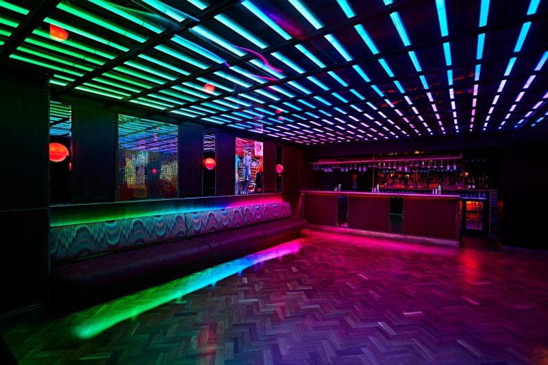 Colorful neon lights and an advanced AV installation illuminate an empty modern bar in London with a vibrant geometric floor pattern, setting a lively yet serene ambiance.