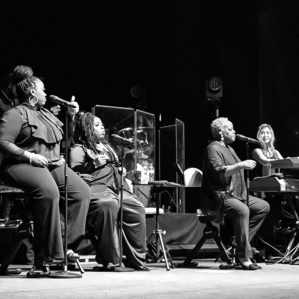 Four musicians in a soulful live performance during their tour production, with three vocalists harmonizing and one pianist accompanying them on stage.