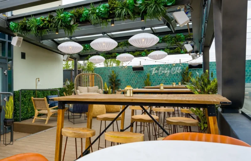 A stylish modern outdoor patio with wooden tables and stools, hanging white lanterns, bar AV installation, and lush greenery, creating an inviting urban oasis for socializing and dining.