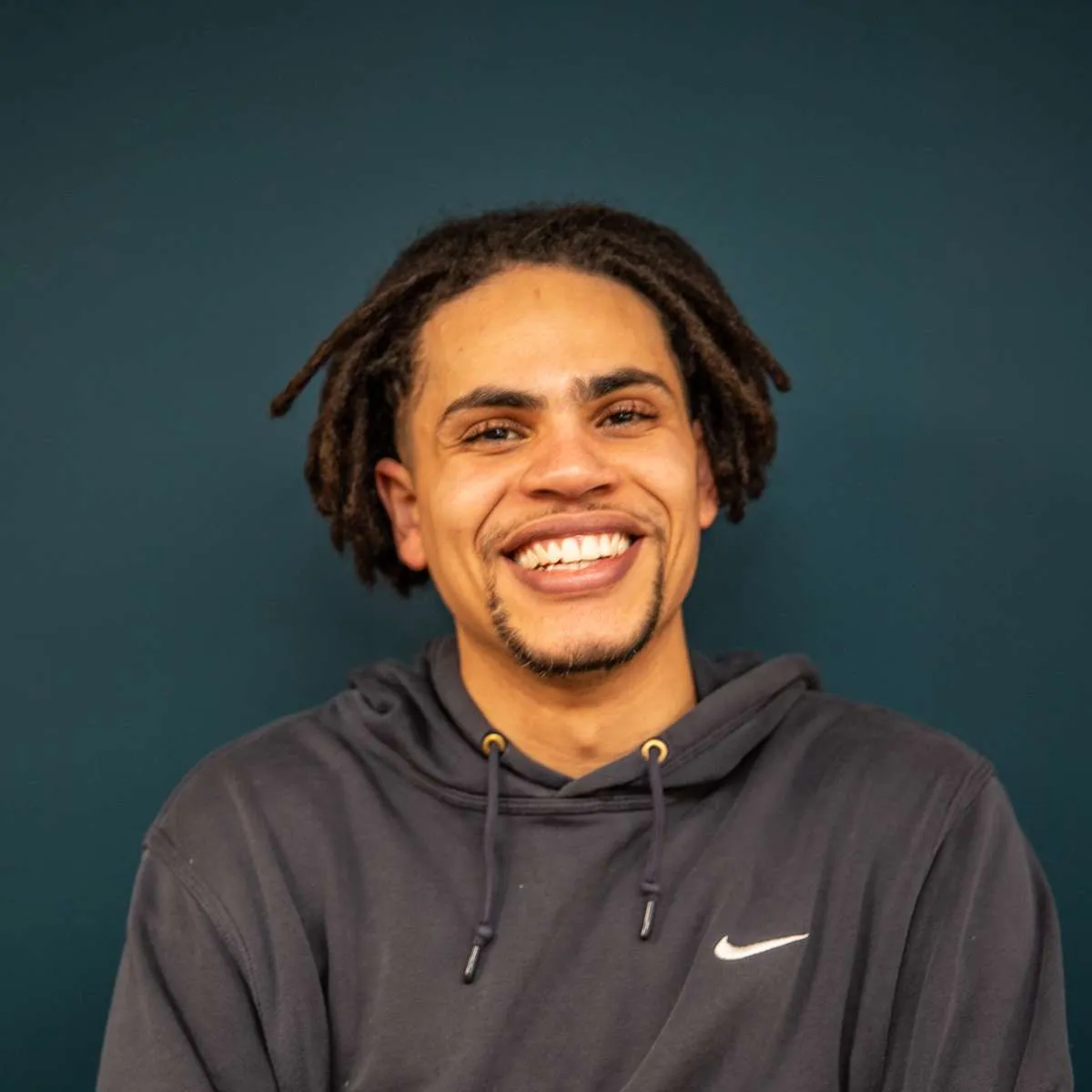 A cheerful young man with dreadlocks smiling broadly in front of a teal background, wearing a dark hoodie with the AV company logo.