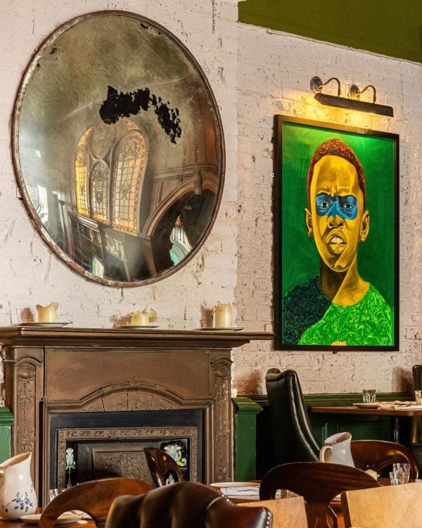 A room with vintage decor featuring an ornate mirror reflecting an interior space designed for a restaurant AV installation, adjacent to a vibrant green painting of a bespectacled individual, all above a classic fireplace