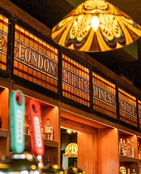A cozy and inviting restaurant atmosphere with warm lighting, featuring a selection of draught beer taps in the foreground and a vintage-style stained glass sign indicating “London spirits wines stout” above wooden shelves.