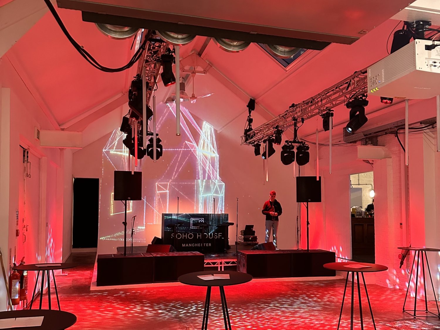 Indoor event space with red lighting and a dj booth labeled "soho dj house manchester". Holographic light structures illuminate the stage, surrounded by empty high-top tables.