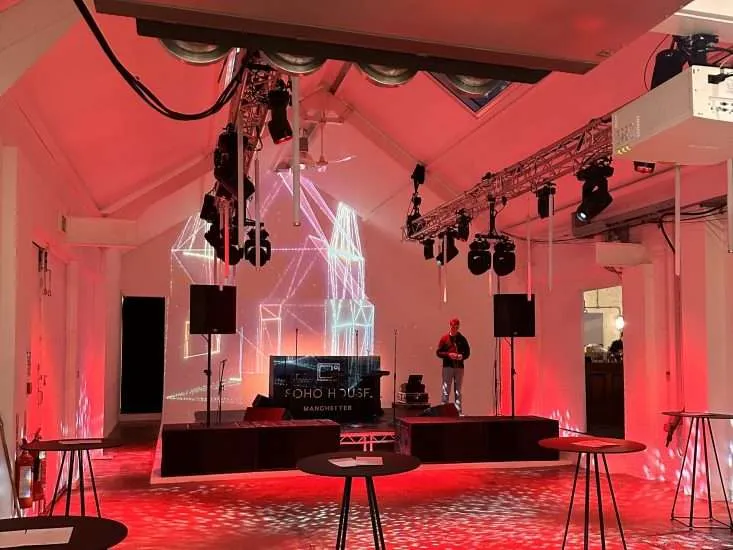 Indoor event space with red lighting and a dj booth labeled "soho dj house manchester". Holographic light structures illuminate the stage, surrounded by empty high-top tables.