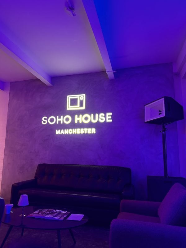 A neon sign reading "soho house manchester" illuminates a room with purple lighting, featuring a dark sofa against a gray wall and an Auto Draft coffee table with magazines.